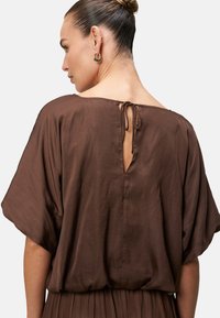 Top marrone a maniche corte con scollatura ampia e chiusura con laccio sul retro. Tessuto morbido e liscio, con un dettaglio centrale a taglio.