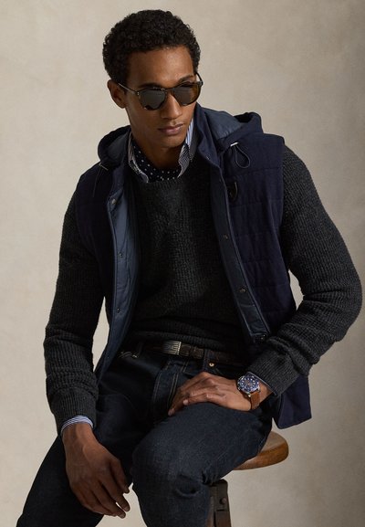 Marineblauw puffer-vest over een donker gebreide trui, gecombineerd met een gestreept overhemd en donkere spijkerbroek. Accessoires zijn een zonnebril en een blauwe horloge.