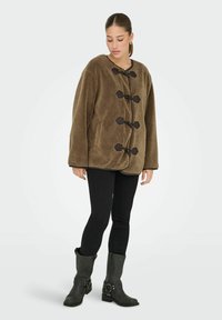 Kobieta nosząca brązowy płaszcz z polarowego materiału z zapięciem na toggle, czarne dopasowane spodnie i czarne kozaki do połowy łydki, stojąca na jednolitym tle.