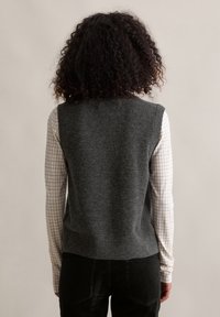 Graue Wollweste mit hohem Kragen, ärmellosem Design und strukturiertem Finish, getragen über einem weiß karierten Hemd.