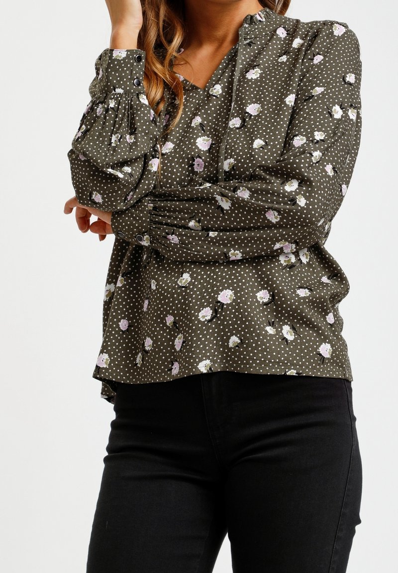 Blouse à manches longues en tissu vert olive à pois blancs et motifs floraux éparpillés, avec un col en V et des poignets boutonnés.