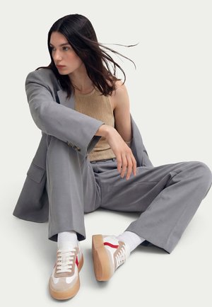 Donna con capelli lunghi e scuri seduta a gambe incrociate, che indossa un completo grigio, una canotta beige senza maniche, calzini bianchi e sneaker bianche con accenti rossi.