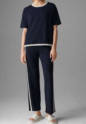 Personne portant un haut bleu marine à manches courtes et un pantalon assorti avec des bandes blanches sur les côtés, associé à des sandales blanches à enfiler.