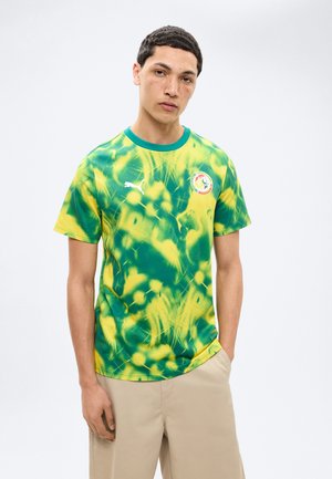 Junger Mann mit kurzen dunklen Haaren, der ein grün-gelbes Tie-Dye-Sporttrikot mit weißem Puma-Logo und beige Hose trägt, vor weißem Hintergrund stehend.