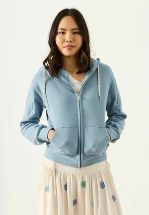 Kvinde med mørkt skulderlangt hår iført en lyseblå hættetrøje med lynlås og et cremefarvet skørt med blå og grøn blomsterbroderi.
