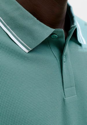 Primo piano di una persona dalla pelle scura che indossa una polo verde con trama, colletto a coste con strisce bianche e grigie.