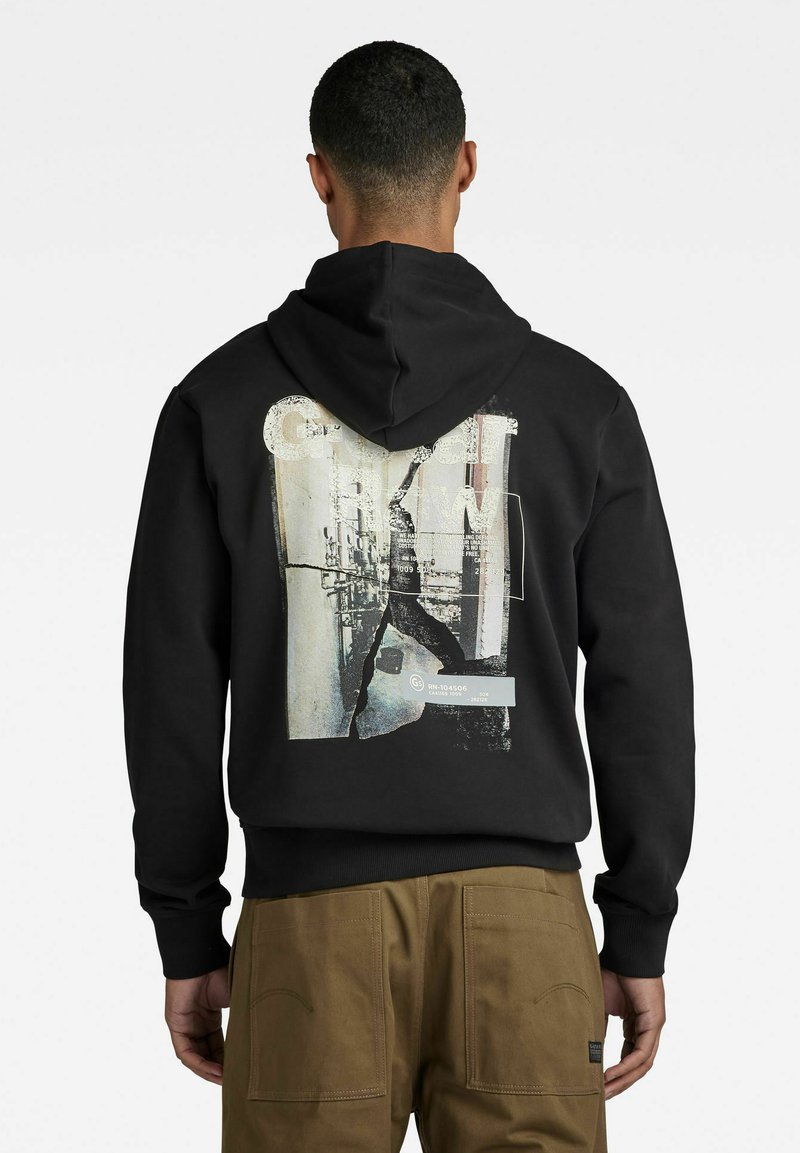 Schwarze Kapuzenjacke mit einem großen, mehrfarbigen Grafikdruck auf der Rückseite, der abstrakte Formen und Text zeigt. Weicher, strukturierter Stoff, normale Passform.