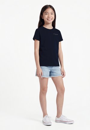 Fille debout, portant une chemise à manches courtes bleu marine, un short en denim clair et des baskets blanches, souriant à la caméra sur un fond blanc.