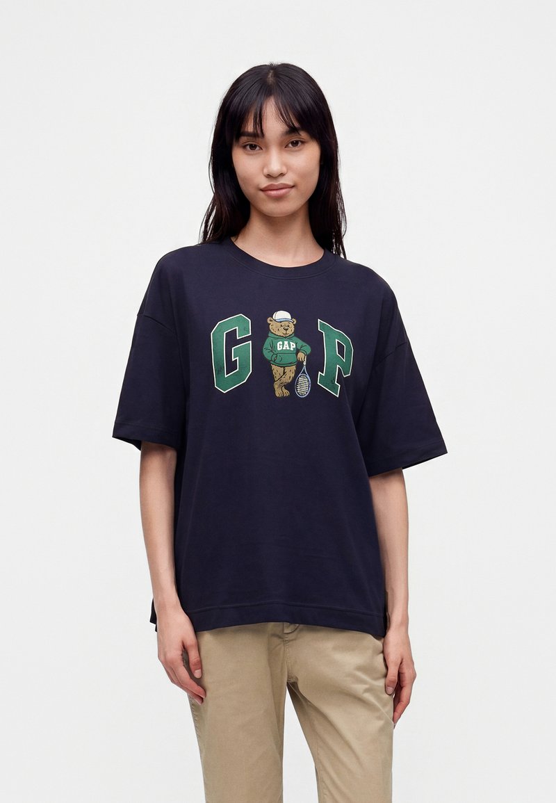 Γυναίκα φορώντας μια μπλε σκούρα oversized μπλούζα με μια πράσινη αρκούδα Gap που κρατά ρακέτα τένις και μπεζ παντελόνι σε απλό φόντο.