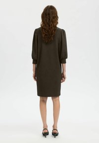 Tummanvihreä, polvipituinen mekko, jossa on liehuvat hihat, sileä pinta ja yksinkertainen, puhdas muotoilu. Mekossa on pyöreä pääntie ja vartalonmyötäinen siluetti.