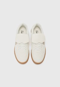 Baskets blanches avec un dessus en cuir texturé, un col rembourré et une sangle à crochet et boucle. La semelle est en caoutchouc marron.