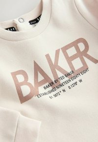 Hellbeige Sweatshirt mit einem großen, metallischen roségoldenen "BAKER"-Druck, zusätzlichem Text darunter und einem Druckknopf am Hals.