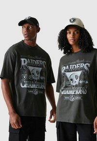 Deux personnes portant des t-shirts graphiques noirs des champions des Raiders du Super Bowl et des casquettes assorties des Raiders, se tenant devant un fond uni.