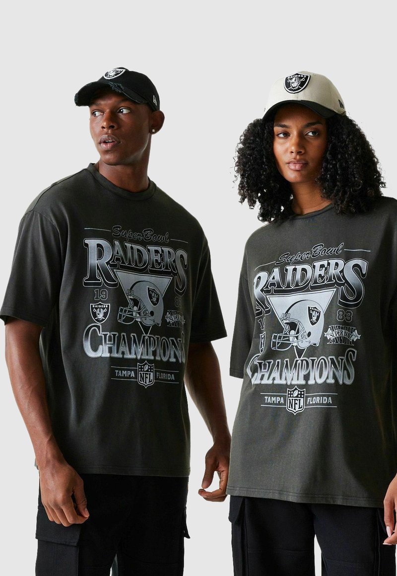 Deux personnes portant des t-shirts graphiques noirs des champions des Raiders du Super Bowl et des casquettes assorties des Raiders, se tenant devant un fond uni.
