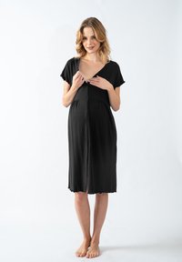 Donna in un vestito nero a bottoni, lungo fino al ginocchio, che sbottona la parte superiore per rivelare l'accesso per l'allattamento, in piedi a piedi nudi su uno sfondo bianco.