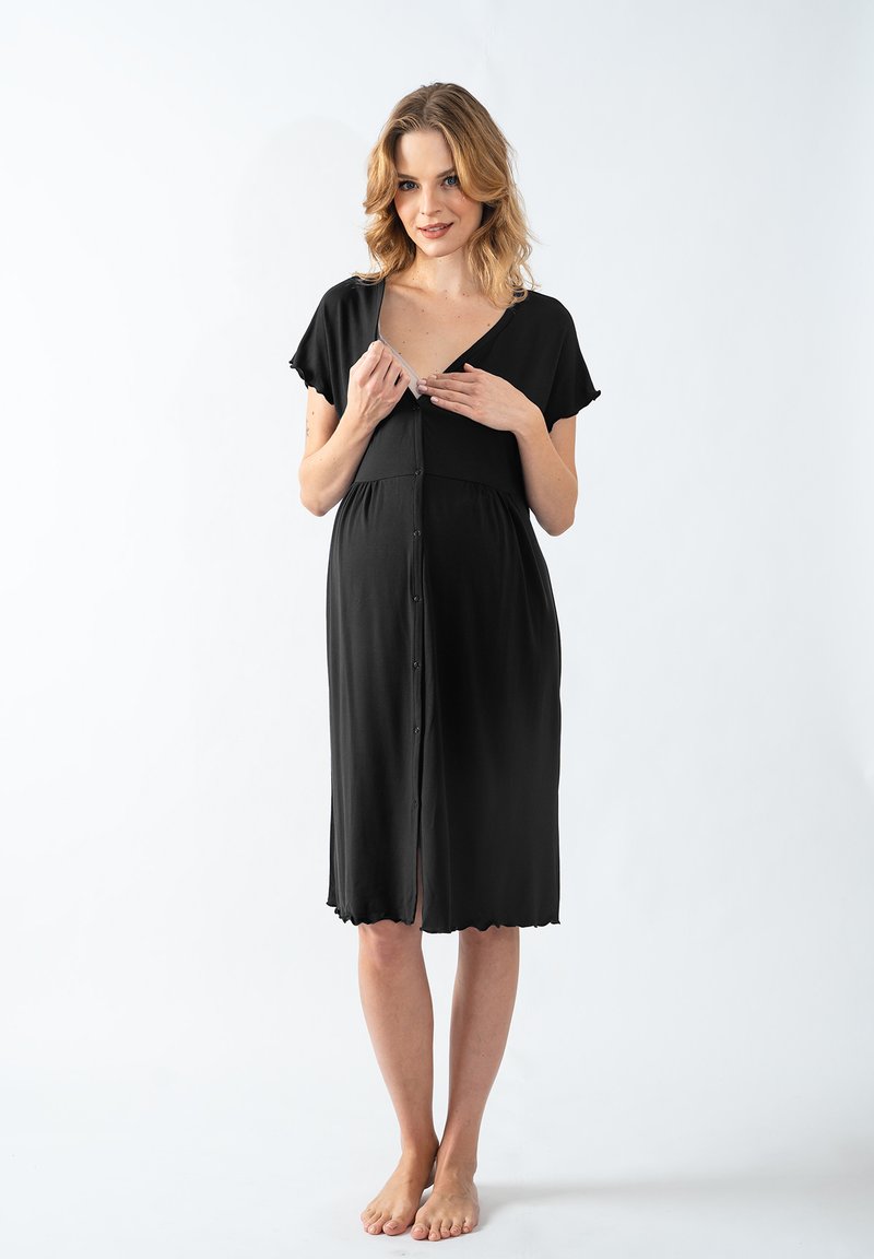 Donna in un vestito nero a bottoni, lungo fino al ginocchio, che sbottona la parte superiore per rivelare l'accesso per l'allattamento, in piedi a piedi nudi su uno sfondo bianco.