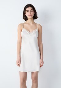 Abito sottoveste in seta bianca lucida con spalline regolabili, bordo in pizzo lungo il collo a V e una silhouette fluida e senza decorazioni.