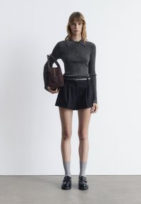 Graues geripptes Langarmoberteil mit Knopfdetails, schwarze plissierte Shorts, graue Socken und schwarze Loafers. Hält eine dunkelbraune Schultertasche.