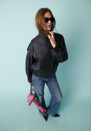 Žena u crnoj kožnoj jakni, plavim trapericama, crnim sunčanim naočalama, držeći malu torbu s crveno-bijelim šalijom na svijetloplatoj pozadini.