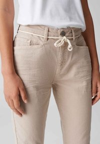 Beige katoenen broek met een trekkoord in de taille, voorzakken en een iets taps toelopende pijp. Zachte textuur met zichtbare stiksel details.