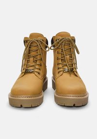 Ein Paar hellbraune Leder-Schnürstiefel mit dicken Sohlen und gemusterten Schnürsenkeln, von vorne auf einem weißen Hintergrund gezeigt.