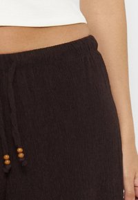 Gros plan d'une personne portant un pantalon texturé foncé avec un cordon orné de perles en bois et un crop top blanc.