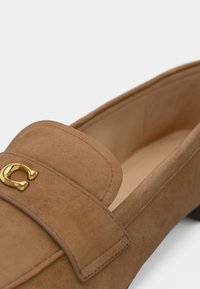 Mocassino in suede tan con hardware dorato, punta leggermente arrotondata e dettagli cuciti; suola piatta e design interno liscio.
