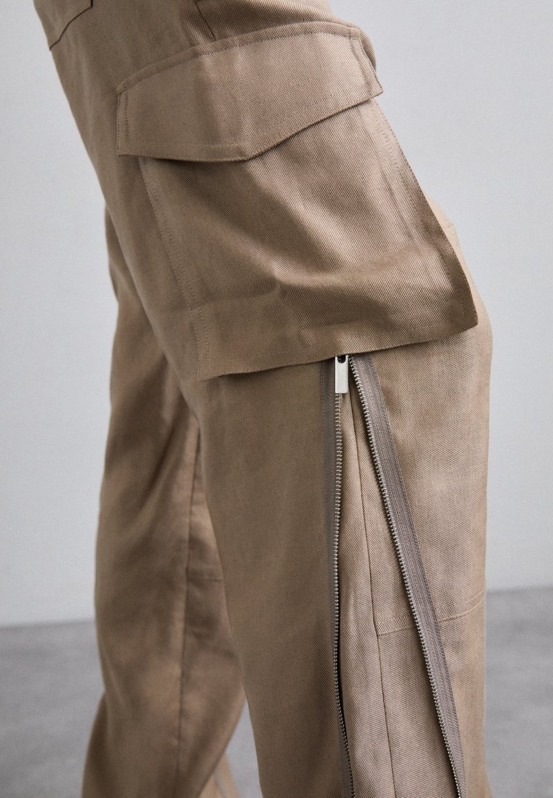 Beige Cargo-Hosen mit seitlichem Reißverschluss, mehreren Taschen, einem strukturierten Stoff und einem lässig lockeren Schnitt, ideal für die Freizeit.