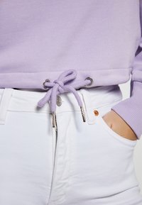Felpa lilla con coulisse e occhielli in argento, abbinata a jeans bianchi e una cerniera visibile. Il tessuto è morbido e dalla consistenza liscia.