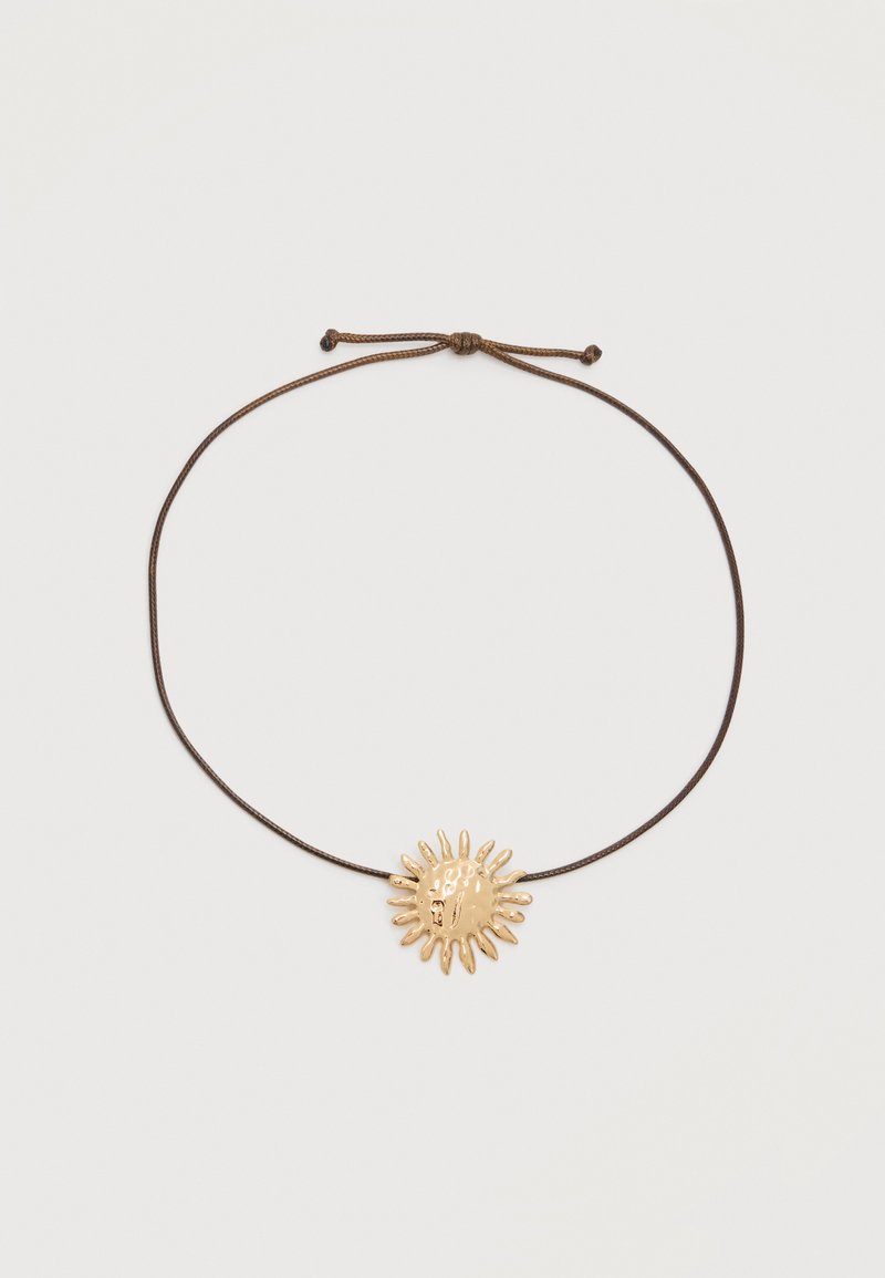 Collier réglable en cordon marron avec un pendentif en forme de soleil doré brillant présentant des rayons irréguliers sur un fond clair.