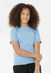 Joven con cabello rizado que lleva una camiseta de Rip Curl de color azul claro, de pie con un brazo sosteniendo el otro contra un fondo blanco.