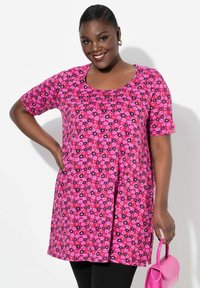 Rosa Blumen-Tunika mit kurzen Ärmeln, verziert mit schwarzen und roten Blumenmustern, kombiniert mit schwarzen Leggings und einer kleinen rosa Handtasche.