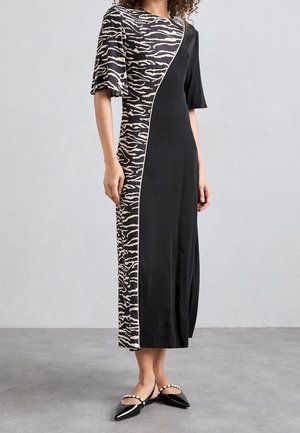 Vrouw met een zwarte midi-jurk met een beige en zwarte zebraprintpaneel en wijd uitlopende mouwen, gecombineerd met zwarte puntige platte schoenen met parelbandjes.