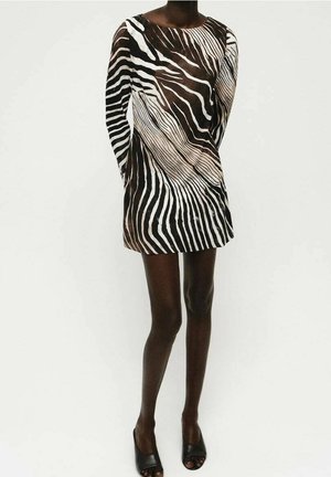 Person, die ein schwarz-weißes Mini-Kleid mit Zebramuster und langen Ärmeln sowie schwarze offene High Heels trägt, vor einem schlichten Hintergrund stehend.