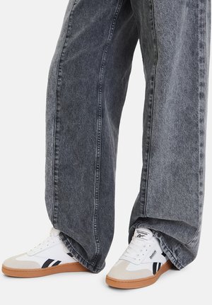 Białe trampki z szarymi akcentami i gumowymi podeszwami, zestawione z luźnymi, ciemnoszarymi spodniami dżinsowymi. Posiadają widoczne przeszycia i szersze nogawki.