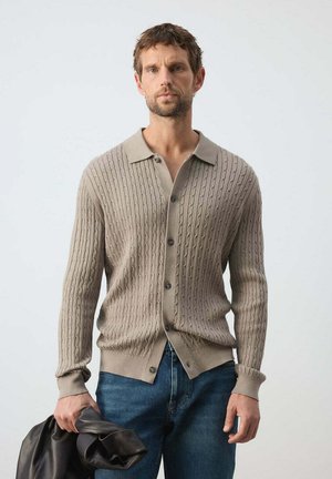Hombre con suéter beige de punto con botones y jeans azules sosteniendo una chaqueta negra de cuero plegada sobre fondo liso.