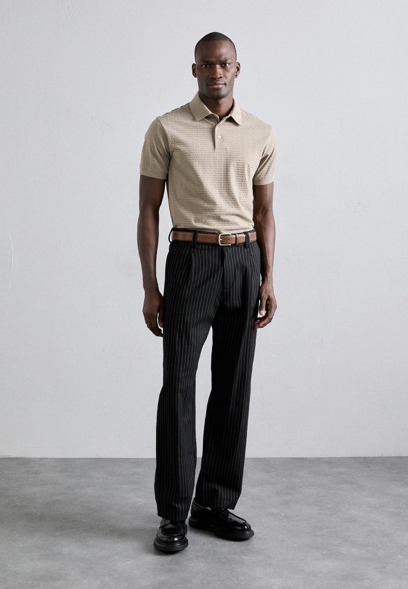 Bežinis raštuotas trumpomis rankovėmis polo marškinėliai, juodi dryžuoti kelniai, rudas diržas, juodi batai. Fone matyti neutralus peizažas su pilku grindų danga.