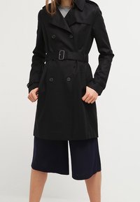 Abrigo negro tipo trench con cierre de botones de doble pecho, cintura ceñida por un cinturón, solapas anchas y textura suave, combinado con culottes azul marino.
