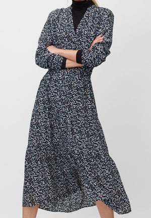 Kvinde iført en lang sort kjole med små hvide og blå blomsterprint, armene krydset, over en sort rullekravebluse.