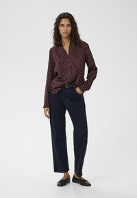 Bluză burgundy cu mâneci lungi și buline albe, cu nasturi, asortată cu blugi înalți, de culoare albastru închis, și pantofi negri fără toc.