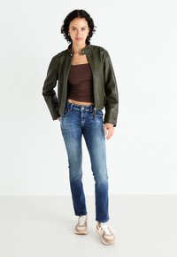 Donkergroene leren jas met een cropped ontwerp, bruine aansluitende top, blauwe spijkerbroek met een lichte vervaging en beige sneakers met witte veters.