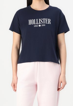 Woman wearing a navy blue cropped T-shirt with "HOLLISTER SINCE 2000" text and light pink pants, standing against a plain background.