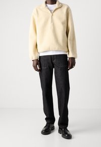 Pullover in pile giallo chiaro con colletto a mezza zip, abbinato a pantaloni di jeans scuri e scarpe nere stringate. Texture liscia e vestibilità rilassata.