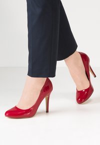 Czerwone lakierowane szpilki z zaokrąglonym noskiem i smukłym obcasem typu stiletto. But posiada błyszczące wykończenie i gładką teksturę.