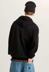 Felpa nera con cappuccio, vestibilità comoda, polsini a coste e vista posteriore. Abbinata a denim azzurro chiaro, con patch logo sulla tasca.