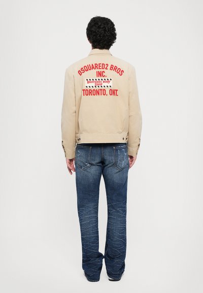 Homme aux cheveux bouclés se tenant de dos, portant une veste beige avec le texte rouge "DSQUARED2 BROS INC. TORONTO, ONT." et un jean bleu.