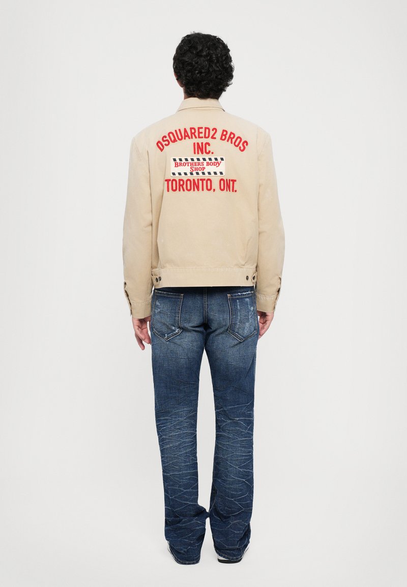 Bărbat cu păr creț stând cu fața îndreptată spre spate, îmbrăcat într-o jachetă bej cu textul roșu "DSQUARED2 BROS INC. TORONTO, ONT." și blugi albaștri.