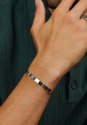 Metalarmbånd med en glat, poleret overflade; har skiftevis flade og strukturerede sektioner; sølvfarvet på et håndled.