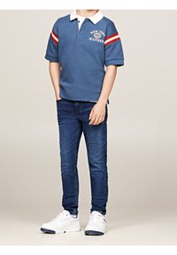 Niebieska koszulka polo z białym kołnierzykiem i czerwonymi paskami, w połączeniu z ciemnoniebieskimi dżinsami i białymi trampkami. Na koszulce znajduje się logo.