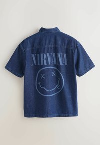 Chemise en denim à manches courtes avec le texte "NIRVANA" et un visage souriant aux yeux barrés et la langue tirée à l'arrière.